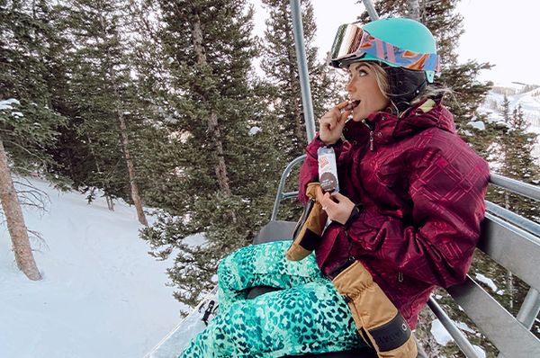 Skifahrerin sitzt in Sessellift und isst einen Riegel.