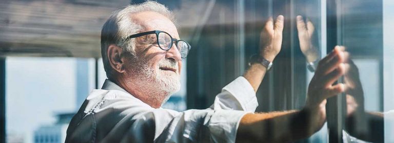 Älterer Mann mit weißen Haaren und Brille steht am Fenster und schaut nach draußen