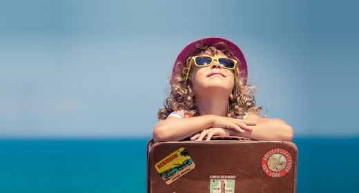 Mädchen mit gelber Sonnebrille genießt die Sonne am Meer und stützt sich auf einen Koffer ab