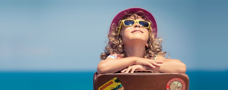 Mädchen mit gelber Sonnebrille genießt die Sonne am Meer und stützt sich auf einen Koffer ab