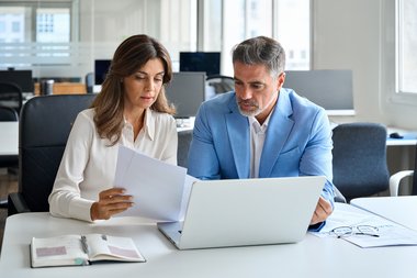 Zwei Personen sitzen im Büro am Laptop und Besprechen eine Unterlage