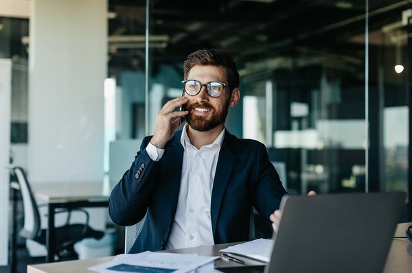 Junger Mann mit Brille sitzt in modernem Büro vor Laptop und telefoniert