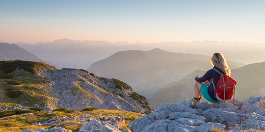 Frau mit Wanderrucksack sitzt am Berggipfel und schaut in den Sonnenuntergang