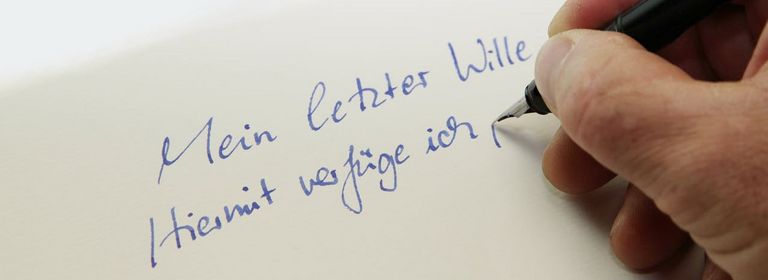 Handschriftliche Worte, geschrieben mit einer blauen Füllfeder auf weißem Papier: Mein letzter Wille. Hiermit verfüge ich ...