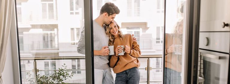 Ein junges Pärchen steht auf dem Balkon und genießen Arm in Arm den Morgenkaffee.