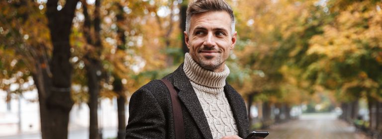 Mann steht in einer herbstlichen Baumallee mit einem Handy und einem Coffee-to-go-Becher in der Hand