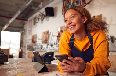 Junge Frau mit orangem Pullover steht in einer Werkstatt und hält ein Handy in der Hand