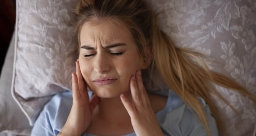 Frau liegt im Bett und hält sich die Wangen vor Schmerz, weil sie um Schlaf mit den Zähnen knirscht