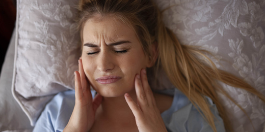 Frau liegt im Bett und hält sich die Wangen vor Schmerz, weil sie um Schlaf mit den Zähnen knirscht