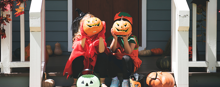 Zwei Kinder in Halloweenkostümen sitzen auf der Veranda vorm Haus und halten sich einen geschnitzten Kürbis vors Gesicht.