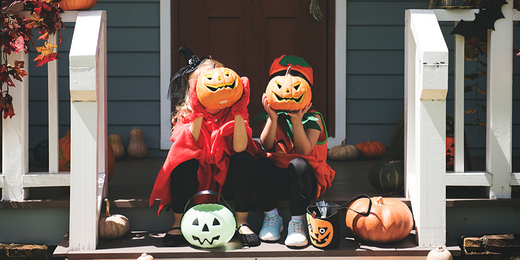 Zwei Kinder in Halloweenkostümen sitzen auf der Veranda vorm Haus und halten sich einen geschnitzten Kürbis vors Gesicht.