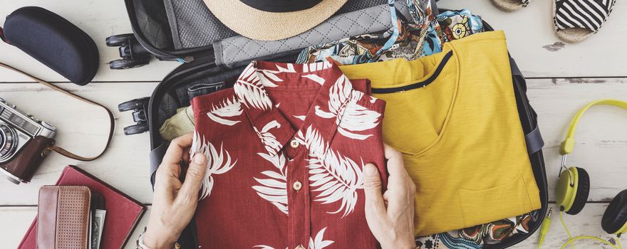 Offener Koffer mit Sommerkleidung liegt am Boden während Person ein Hemd hineinlegt