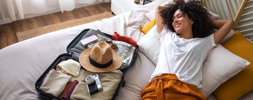 Frau liegt entspannt auf dem Bett neben ihrem gepackten Koffer und freut sich, dass ihr die Checkliste für den Urlaub Stress erspart hat. 